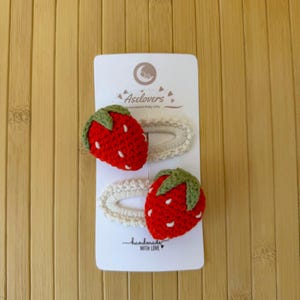 May include: Two crochet hair clips on a white display card labeled Aselovers. Each clip features a red strawberry with a green stem and white embroidered seeds attached to an oval-shaped, off-white crocheted base. The card includes the text handmade WITH LOVE.