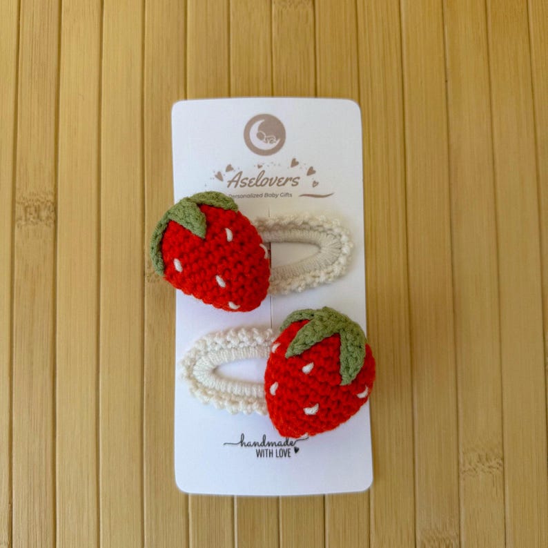 May include: Two crochet hair clips on a white display card labeled Aselovers. Each clip features a red strawberry with a green stem and white embroidered seeds attached to an oval-shaped, off-white crocheted base. The card includes the text handmade WITH LOVE.