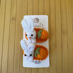 May include: Two crochet hair clips attached to a white display card. Each clip features a white rabbit with black eyes and a red stitched mouth, positioned next to an orange carrot with green leaves. The card displays the text "elovers" and "handmade WITH LOVE".