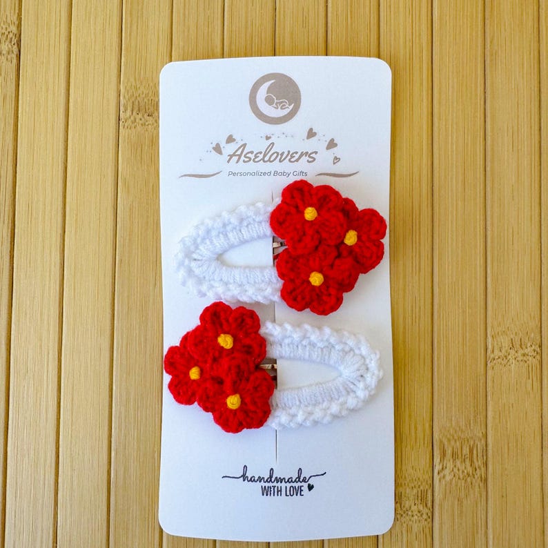 May include: Two oval hair clips featuring white crocheted borders and clusters of three bright red flowers with yellow centers. The clips are attached to a white display card labeled Aselovers Personalized Baby Gifts and handmade with love.