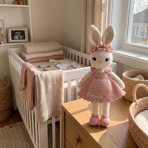 K&ouml;nnte beinhalten: Eine handgefertigte H&auml;kelkaninchenpuppe in einem rosa Kleid mit dem Namen "EMMA" aufgestickt. Die Puppe hat lange Ohren und steht auf einer Holzoberfl&auml;che. Im Hintergrund sind ein Kinderbett, Decken und ein Fenster zu sehen.