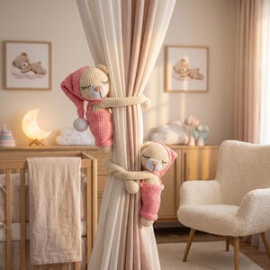 May include: Two cream-coloured crocheted teddy bears with closed eyes, each wearing a pink nightcap and pink overalls. The bears are positioned to hold onto a set of light-coloured curtains, with one bear placed higher than the other on the fabric.