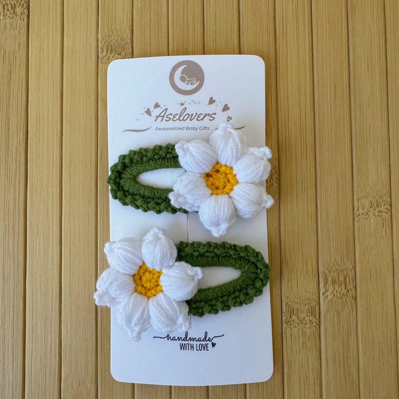 May include: Two crochet hair clips on a white display card labeled Aselovers Personalized Baby Gifts and handmade WITH LOVE. Each clip features a green oval base with a white flower with a yellow center attached to the top of the crochet fabric.