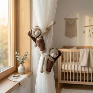 May include: Two crocheted bear-shaped curtain tiebacks in shades of beige and brown. Each bear features a sleeping expression with closed eyes, a small snout, and long arms designed to wrap around and secure a sheer white curtain panel.