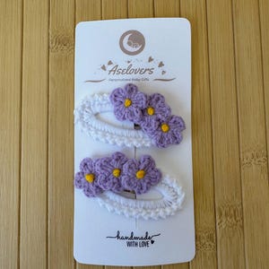 May include: Two oval-shaped hair clips featuring white crocheted borders and three light purple crocheted flowers with yellow centers. The clips are attached to a white display card labeled Aselovers Personalized Baby Gifts and handmade with love.