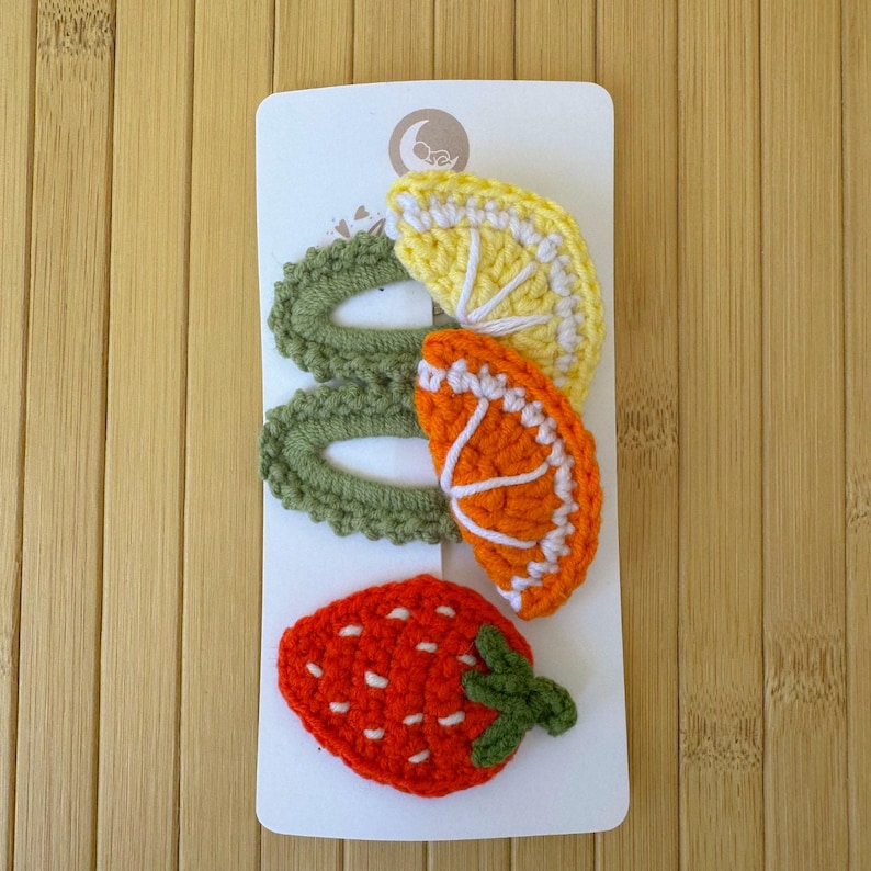 May include: A set of three crochet hair clips on a white display card. Two clips feature green oval bases topped with yellow lemon and orange citrus slice motifs. The third clip is a red strawberry with green leaves and white seed details.
