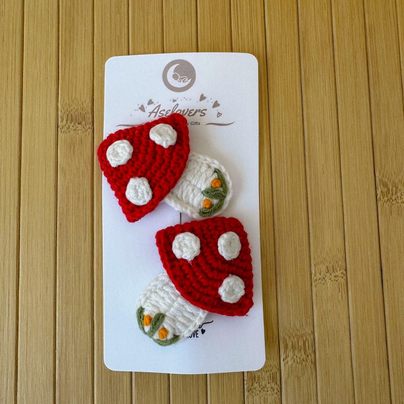 May include: Two crochet mushroom hair clips attached to a white display card with the text Aselovers. Each mushroom features a bright red cap with white spots and a white stem decorated with small green leaves and tiny orange accents.
