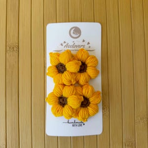 May include: Two crocheted sunflower hair clips attached to a white display card. The flowers feature bright yellow yarn petals and dark brown centers. The card displays the text Aselovers and handmade WITH LOVE with a small heart icon.