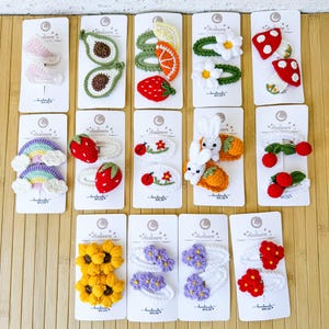 May include: A collection of fourteen sets of handmade crochet hair clips attached to white display cards. The designs include avocados, citrus slices, strawberries, daisies, mushrooms, rainbows, ladybugs, rabbits, cherries, sunflowers, and purple and red flowers.