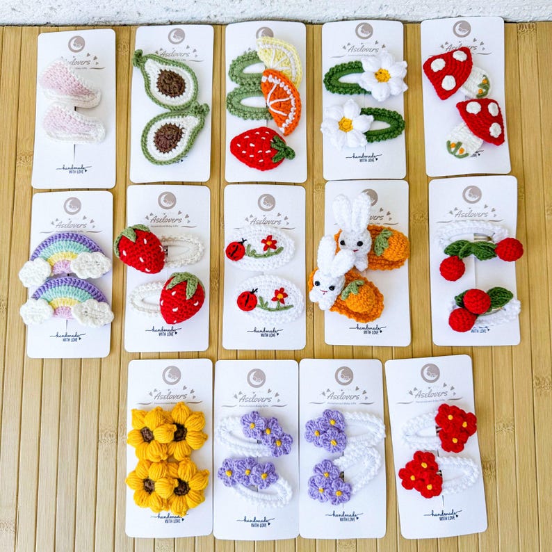 May include: A collection of fourteen sets of handmade crochet hair clips attached to white display cards. The designs include avocados, citrus slices, strawberries, daisies, mushrooms, rainbows, ladybugs, rabbits, cherries, sunflowers, and purple and red flowers.