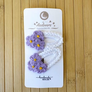 May include: Two white crochet hair clips on a display card labeled Aselovers Personalized Baby Gifts and handmade WITH LOVE. Each clip features a cluster of three light purple crocheted flowers with small yellow centers attached to a white base.