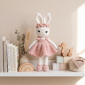 K&ouml;nnte beinhalten: Eine handgefertigte, wei&szlig;e H&auml;kelkaninchenpuppe mit rosa Kleid und passenden Schuhen, mit einem Blumenstirnband. Die Puppe steht auf einem Regal mit Holzspielzeug, B&uuml;chern und einem kleinen Korb. Die Puppe ist etwa 38 cm gro&szlig;.