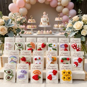 May include: A collection of crochet hair clips displayed on individual cards labeled Emma's Baby Shower. The clips feature various designs including fruits like strawberries, oranges, and avocados, as well as flowers, rainbows, and a small white bunny.