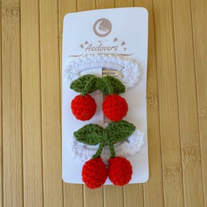 May include: Two hair clips on a white display card labeled Aselovers. Each clip features a white crocheted base topped with two bright red crocheted cherries and two green crocheted leaves attached to a small metal snap clip mechanism.