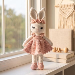 K&ouml;nnte beinhalten: Handgefertigte H&auml;kelkaninchenpuppe mit hellrosa Kleid, passenden Schuhen und einem Blumenstirnband. Das H&auml;schen hat lange Ohren und schwarze Augen. Es steht auf einer wei&szlig;en Oberfl&auml;che in der N&auml;he eines Fensters.