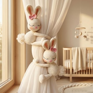 May include: Two cream-coloured crocheted rabbit curtain tiebacks featuring pink inner ears, small pink bows, and fluffy white tails. Each rabbit has closed eyes and a pink nose, with long arms wrapped around a sheer white curtain panel.