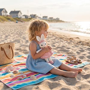K&ouml;nnte beinhalten: Ein Kind sitzt auf einem gestreiften Strandtuch und h&auml;lt eine handgefertigte wei&szlig;e und rosa Hasenpuppe. Die Puppe hat ein rosa Kleid mit dem Namen "EMMA". Eine Strohtasche und Sandalen liegen in der N&auml;he.