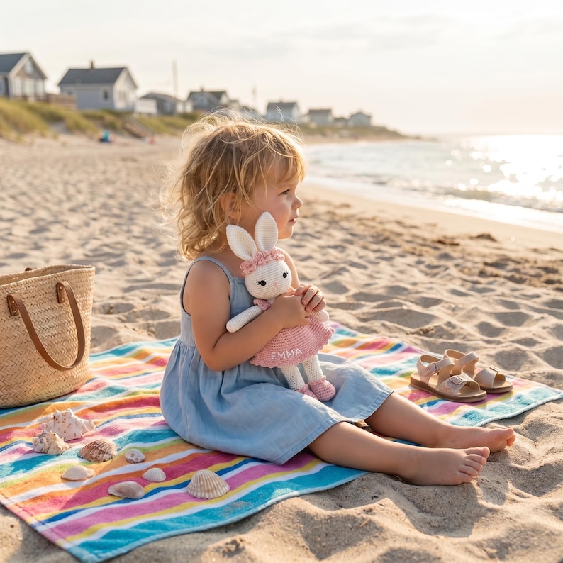 K&ouml;nnte beinhalten: Ein Kind sitzt auf einem gestreiften Strandtuch und h&auml;lt eine handgefertigte wei&szlig;e und rosa Hasenpuppe. Die Puppe hat ein rosa Kleid mit dem Namen "EMMA". Eine Strohtasche und Sandalen liegen in der N&auml;he.