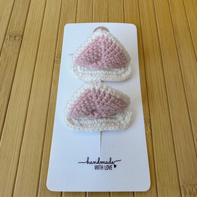 May include: Two triangular crochet cat ear hair clips featuring a soft pink center and a white border. The items are attached to a white rectangular display card with the text handmade WITH LOVE printed in black at the bottom center.