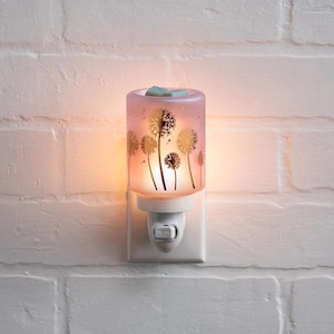 Puede incluir: Luz de noche decorativa con una pantalla de cristal rosa con un diseño de diente de león. La luz de noche está enchufada a una toma de corriente blanca y está iluminada, emitiendo un brillo cálido. El diseño del diente de león es dorado.