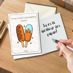May include: Greeting card with the text "Tu es le meilleur des papas!" and a cartoon corn dog illustration. The card is on a wooden surface with pens, an envelope, and a gift box in the background.
