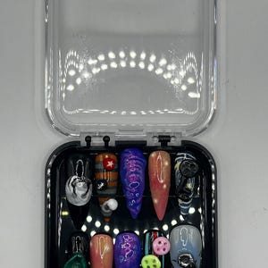 Könnte beinhalten: Ein Set aus zehn künstlichen Nägeln in einer transparenten Kunststoffbox. Die Nägel weisen verschiedene Designs auf, darunter ein Spinnennetz, ein Stern und abstrakte Muster in Blau-, Rosa- und Grüntönen. Die Nägel sind in unterschiedlichen Längen und Formen.