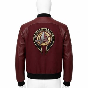 May include: A maroon bomber jacket with black ribbed collar, cuffs, and hem. The back features a large, circular embroidered patch with the text STARFLEET ACADEMY surrounding a central metallic-coloured delta insignia on a black and gold background.