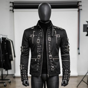 May include: Black jacket with multiple silver buckles and zippers. The jacket has a high collar and is displayed on a mannequin. The sleeves have silver rivets and the jacket is made of a smooth, possibly leather-like material.