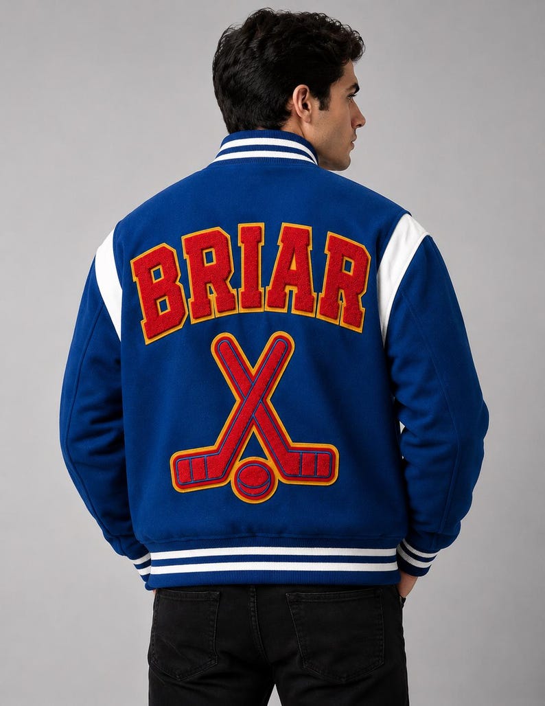 May include: A blue varsity jacket with white sleeves and trim. The back features the word "BRIAR" in red letters above a hockey stick and puck design. The jacket has a ribbed collar and cuffs.
