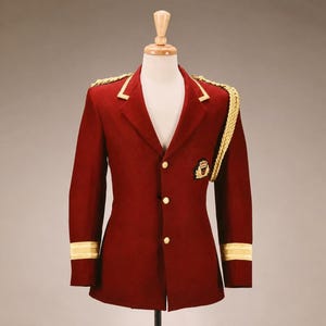 Op de afbeelding: Een bordeauxrode blazer met gouden details, gepresenteerd op een mannequin. De jas heeft gouden biezen op de kraag en manchetten, plus gouden epauletten en knopen. Een klein embleem staat op de borst.