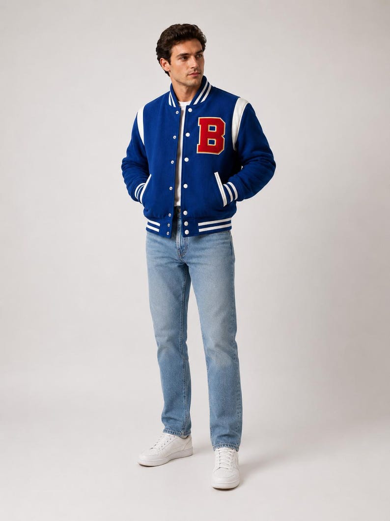 May include: A blue varsity jacket with white accents and a large red "B" on the chest. The jacket has a button-up front and white stripes on the cuffs and hem. The person is wearing light blue jeans and white sneakers.