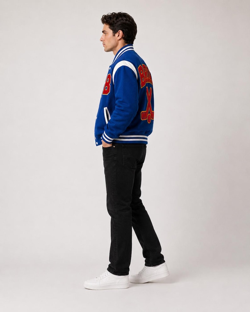 May include: A blue varsity jacket with white and red accents. The jacket features the word "Bruins" and crossed hockey sticks on the back. The jacket has a button-up front, white stripes on the cuffs and hem, and a white collar.