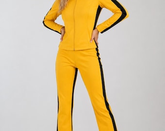 Op de afbeelding: Een felgeel trainingspak bestaande uit een jack met ritssluiting en opstaande kraag en een lange broek. Zowel het jack als de broek zijn voorzien van opvallende zwarte verticale strepen langs de buitenkant van de armen en de benen.