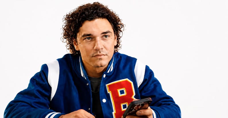 May include: A blue and white varsity jacket with a large red "B" patch. The jacket is worn by a person holding a smartphone. The person has curly brown hair and is looking off to the side.