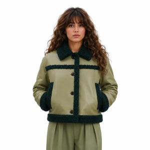 May include: An olive green jacket with a dark green faux fur collar, trim, and cuffs. The jacket has a button-up front and two pockets. The model is wearing the jacket. The jacket is a stylish outerwear piece.