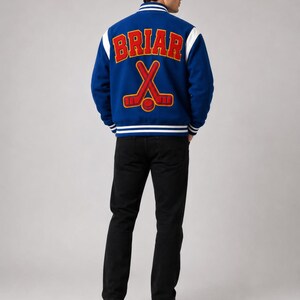 May include: A blue varsity jacket with white sleeves and trim. The back of the jacket features the word "BRIAR" in red block letters above a crossed hockey stick design. The person is wearing black jeans and white sneakers.