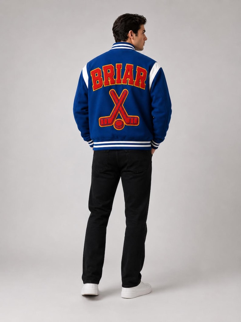 May include: A blue varsity jacket with white sleeves and trim. The back of the jacket features the word "BRIAR" in red block letters above a crossed hockey stick design. The person is wearing black jeans and white sneakers.