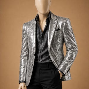 Può includere: Un blazer argentato con paillettes, una camicia nera e pantaloni neri. Il blazer ha una chiusura a due bottoni e un fazzoletto da taschino. La camicia ha un sottile motivo a righe. Il manichino è su sfondo marrone.