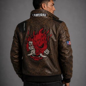 Puede incluir: Una chaqueta de cuero marrón con aspecto desgastado que presenta un gran gráfico bordado de una máscara oni roja y blanca en la espalda. El cuello muestra el texto SAMURAI en blanco, y un parche negro en el hombro dice CRYPTALL JOCK.