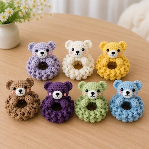Puede incluir: Una colección de siete anillos de ganchillo con forma de oso en varios colores, incluyendo lavanda, crema, amarillo, marrón, morado oscuro, verde claro y azul claro. Cada oso tiene ojos negros pequeños, nariz bordada y mejillas rosas.