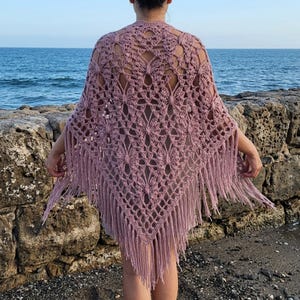 Può includere: Uno scialle all'uncinetto color rosa polvere caratterizzato da un intricato motivo traforato ispirato alle farfalle. Il capo triangolare è rifinito con lunghe frange pendenti lungo i bordi inferiori, creando un aspetto strutturato.