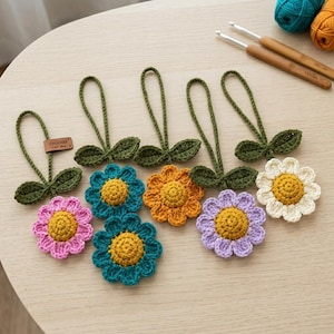 May include: Six crocheted flower ornaments with yellow centers and petals in pink, teal, orange, lavender, and white. Each flower features a green stem with two leaves and a loop for hanging. A small brown tag labeled CROCHIEF is attached to one.