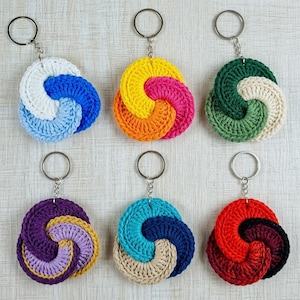 May include: Six circular crochet keychains arranged in two rows. Each keychain features a metal ring attached to a three-colored, spiraled yarn design. The colors include combinations of white, blue, yellow, orange, pink, green, purple, teal, beige, red, and black.