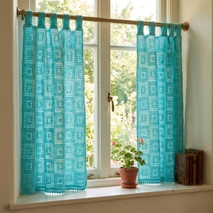 Op de afbeelding: Turquoise gehaakte gordijnen met een geometrisch patroon hangen voor een raam. De gordijnen zijn opgehangen aan een goudkleurige stang. Een kleine plant in een pot staat op de vensterbank, samen met een paar boeken.