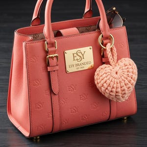 May include: A coral-colored handbag with embossed "ESY" lettering and gold hardware. The bag features a gold plaque with "ESY BRANDED EST. 2024". A crocheted heart charm hangs from the handle. Sunglasses rest inside the open bag.