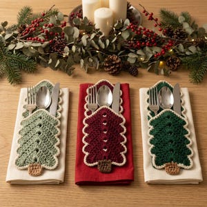 Peut inclure: Trois ensembles de couverts sont glissés dans des pochettes en crochet en forme de sapin de Noël. Les pochettes sont vert clair, rouge foncé et vert foncé, avec un tronc marron et une bordure crème, posées sur des serviettes beige et rouge.