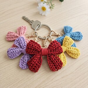 Può includere: Una collezione di portachiavi a fiocco fatti a mano all'uncinetto in vari colori, tra cui rosso, viola, rosa, giallo e blu. Ogni fiocco è attaccato a un fermaglio color oro e a un portachiavi argentato. È presente anche una chiave.