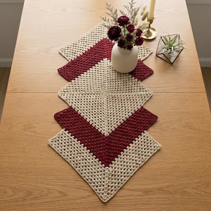 May include: A crochet table runner featuring a geometric pattern of alternating cream and deep red square motifs. The runner is laid out in a diamond orientation, showcasing a textured, open-work stitch design throughout the entire piece.