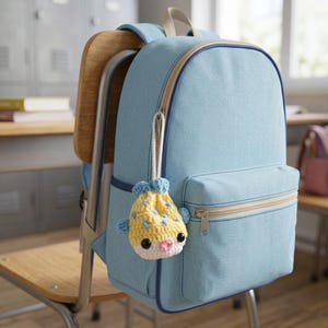 May include: A light blue backpack with a front zippered pocket and a top handle. Hanging from the zipper is a small, crocheted pufferfish charm in yellow, white, and blue with black eyes and a pink mouth, attached by a white cord loop.