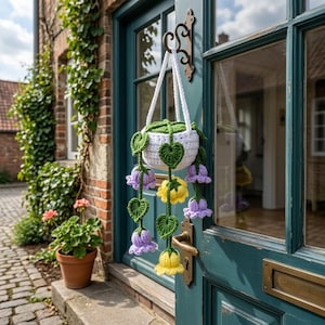 May include: A white crochet hanging planter with green leaves and purple and yellow flower accents. The planter is suspended from a decorative metal hook on a teal door. The background includes a brick building and a potted plant.